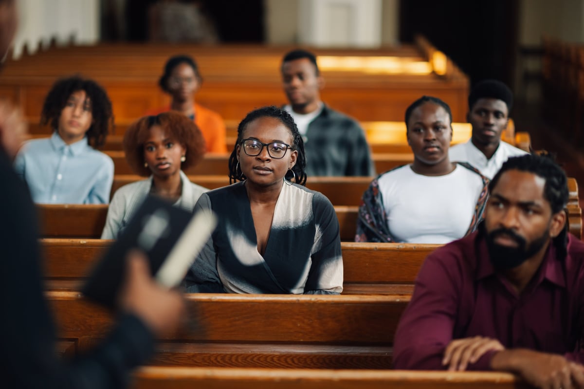 Diverse churchgoers sit in wooden pews, listening intently to the preacher's sermon